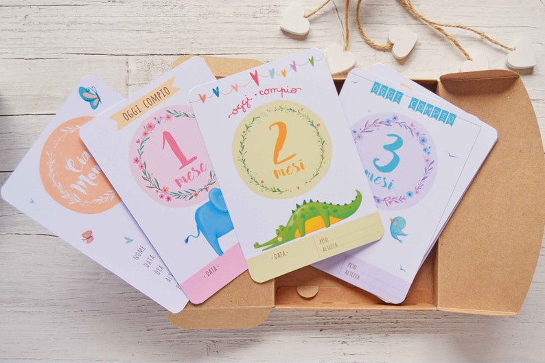 May include: Four colorful baby milestone cards with illustrations of an elephant, crocodile, and bird. The cards are in a cardboard box and feature the text "Oggi compio" and numbers 1, 2, and 3.