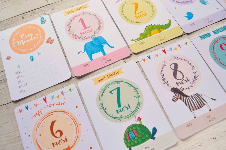 May include: A set of ten baby milestone cards with colorful illustrations of animals and the text "Oggi compio" (meaning "Today I turn") and the number of months. The cards are designed for a baby's first year.