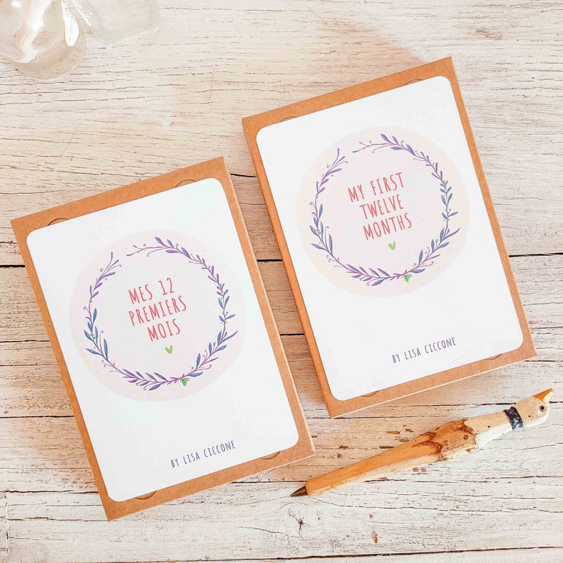May include: Two brown boxes with white covers. The covers feature a pink circle with a floral wreath and text. The left box says "Mes 12 Premiers Mois" and the right box says "My First Twelve Months". Both boxes have the text "By Lisa Ciccone" at the bottom.