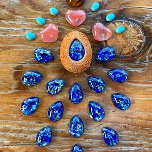 May include: An assortment of gemstones and carved wooden pieces arranged on a wooden surface. The collection includes blue, turquoise, and pink stones, along with a carved wooden oval with a blue stone center. A tiger's eye stone is also present.