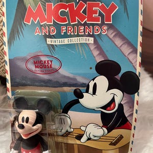 May include: Mickey Mouse and Friends Vintage Collection action figure. The packaging features Mickey Mouse playing a guitar, with a tropical background. The figure is in a clear plastic case. The packaging has the text "Mickey Mouse Action Figure".