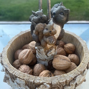 May include: A decorative nutcracker featuring three squirrels on a wooden bowl filled with walnuts. The squirrels are in shades of grey and brown, with a silver nutcracker mechanism. The bowl has a textured, bark-like appearance.