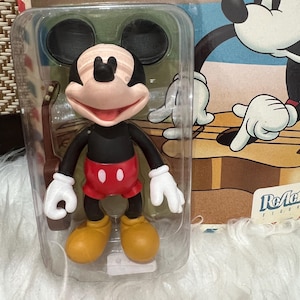 May include: A Mickey Mouse action figure in a clear plastic package. The figure has a black body, red shorts, yellow shoes, and white gloves. The packaging features a cartoon image of Mickey Mouse playing a guitar.