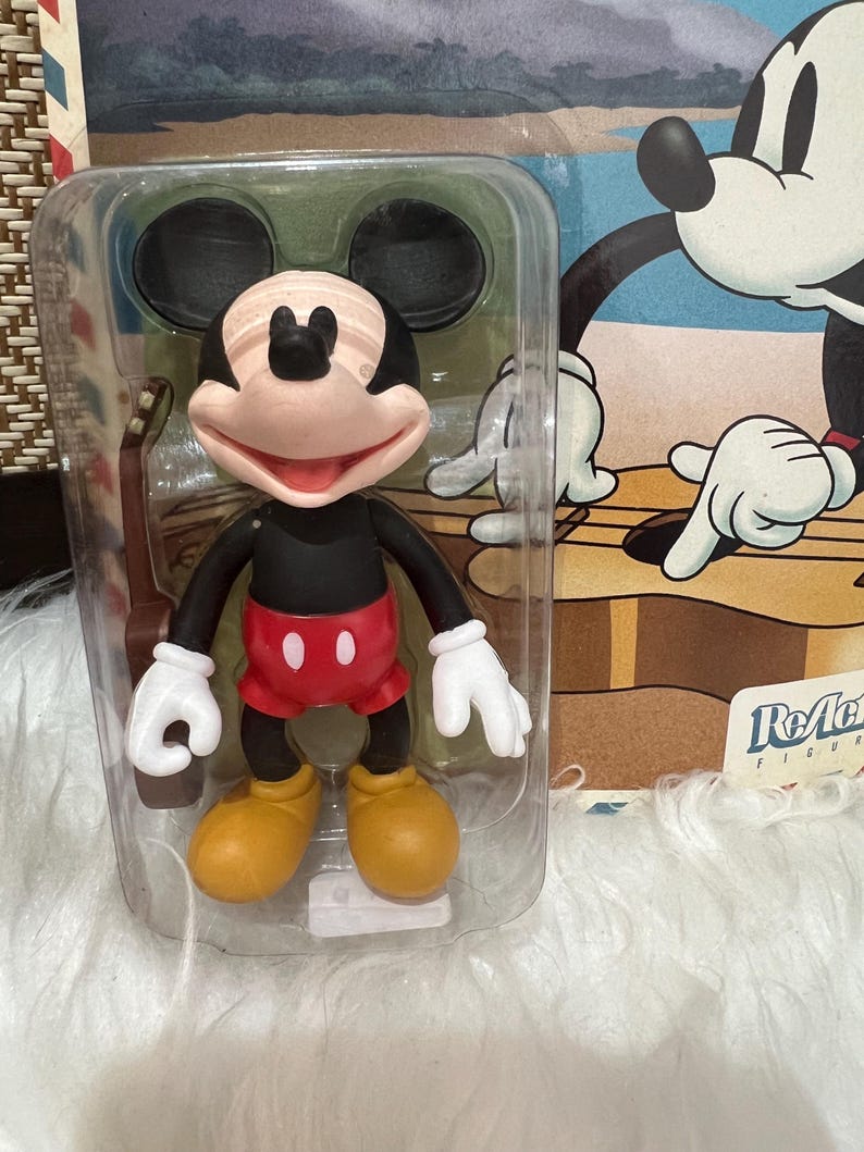 May include: A Mickey Mouse action figure in a clear plastic package. The figure has a black body, red shorts, yellow shoes, and white gloves. The packaging features a cartoon image of Mickey Mouse playing a guitar.