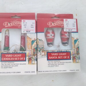 May include: Two Christmas yard light sets in boxes. One set features red candles with "NOEL" text, the other features Santa figures. Both sets are battery-operated and made exclusively for Lemax.