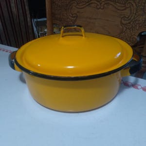 May include: A yellow enamel pot with a matching lid. The pot has black accents on the handles and rim. The pot is sitting on a white surface.