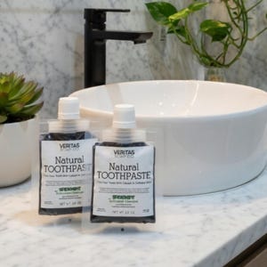 Puede incluir: Dos botellas de pasta de dientes natural Veritas Soap Co. con carbón activado y colágeno, expuestas sobre una encimera de mármol junto a un lavabo blanco y una pequeña planta suculenta en una maceta blanca. El texto de las botellas dice "Natural Toothpaste".