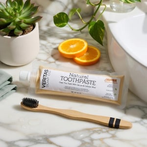 May include: A natural toothpaste tube and a bamboo toothbrush are displayed on a marble surface. The toothpaste tube has the text "Natural Toothpaste" and the brand name "Veritas Soap Co." The toothbrush has black stripes. Two orange slices and a succulent plant in a white pot are also visible.