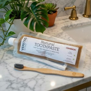 Puede incluir: Un tubo de "Natural Toothpaste" con sabor a "Cinnamon", un cepillo de dientes de bambú y un paño blanco se exhiben en una encimera de mármol. El tubo de pasta de dientes es transparente, mostrando la pasta marrón en su interior. El cepillo de dientes tiene cerdas grises.