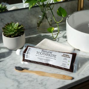 Puede incluir: Un tubo de "Natural Toothpaste Double Chocolate" y un cepillo de dientes de madera sobre una encimera de baño. El tubo de pasta de dientes es marrón oscuro con texto blanco. Una pequeña planta suculenta y un jarrón de cristal también son visibles.