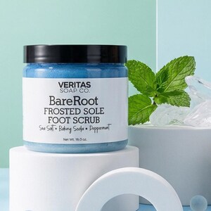 Könnte beinhalten: Ein Glas BareRoot Frosted Sole Fußpeeling von Veritas Soap Co. wird mit Minzblättern und Eis gezeigt. Das blaue Peeling wird mit einem Löffel entnommen. Der Text auf dem Bild lautet "Die Kur für müde, schmerzende Füße."
