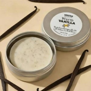 May include: A skincare product with the text "MEET YOUR NEW SKIN ESSENTIAL". The image features a round, light-coloured soap bar in a silver tin, with the words "Warm Vanilla" on the lid. Several dark brown vanilla beans are scattered around the tin.