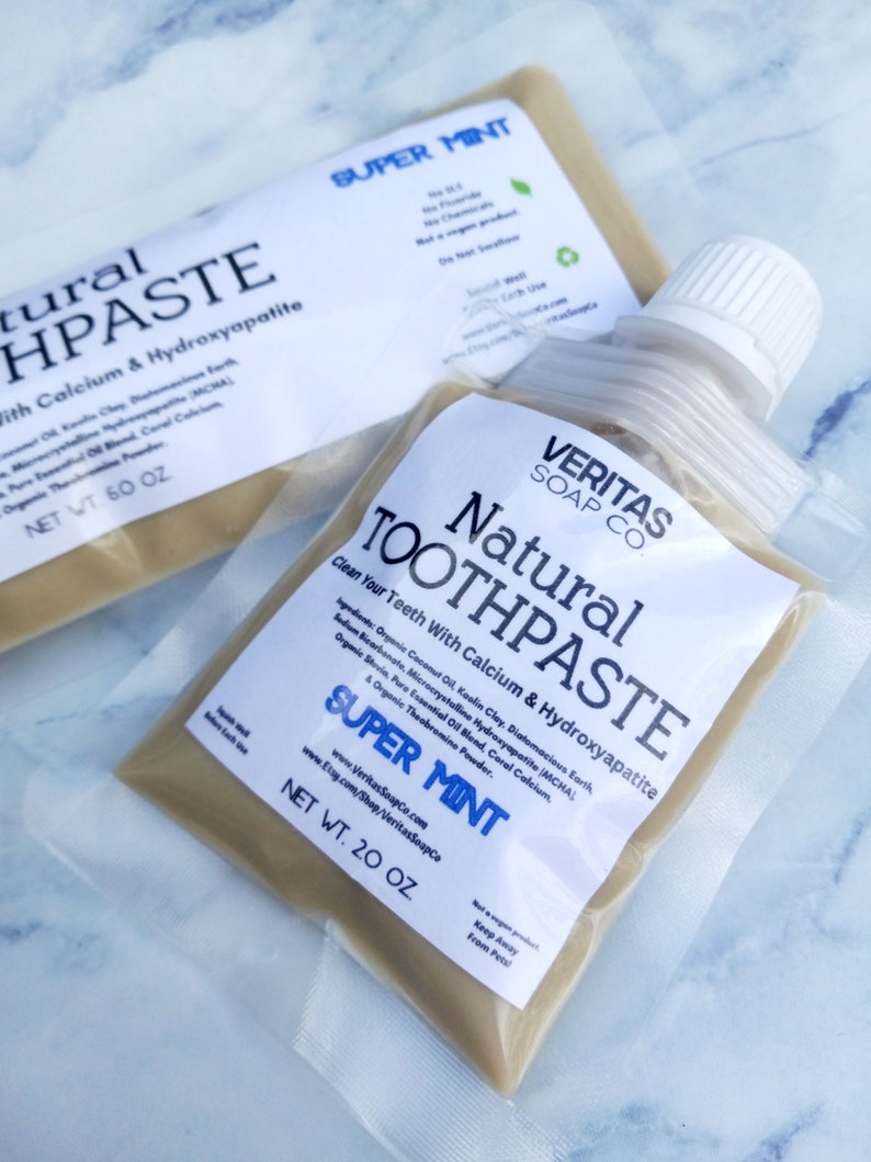 Op de afbeelding: Twee zakjes natuurlijke tandpasta met de tekst "SUPER MINT" en "Natural Toothpaste" in een doorzichtig plastic zakje. De tandpasta is lichtbeige van kleur. Het nettogewicht is 567 g en 1701 g.