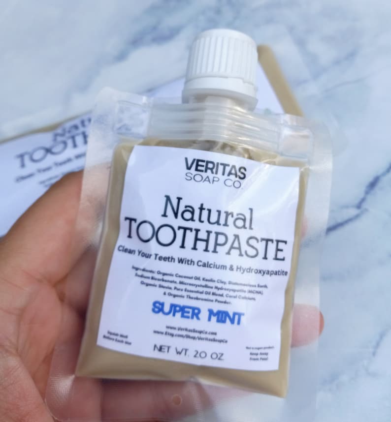 Op de afbeelding: Een doorzichtige zak van "Natural Toothpaste" van Veritas Soap Co. Het etiket luidt "Super Mint" en "Net Wt. 56,7 g." De tandpasta is lichtbeige van kleur. De zak heeft een witte dop.