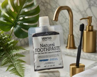 May include: A package of Veritas Soap Co. Natural Toothpaste, with the text "Peppermint", sits on a marble surface. A black toothbrush in a gold holder, a gold soap dispenser, and a gold faucet are also visible. The toothpaste is 2.0 oz.