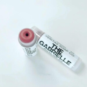 May include: Two lip balm tubes are shown on a white surface. One is open, displaying a pink balm with a dark centre. The other is closed, with the text "VERITAS SOAP CO. THE GABRIELLE" printed on the label.