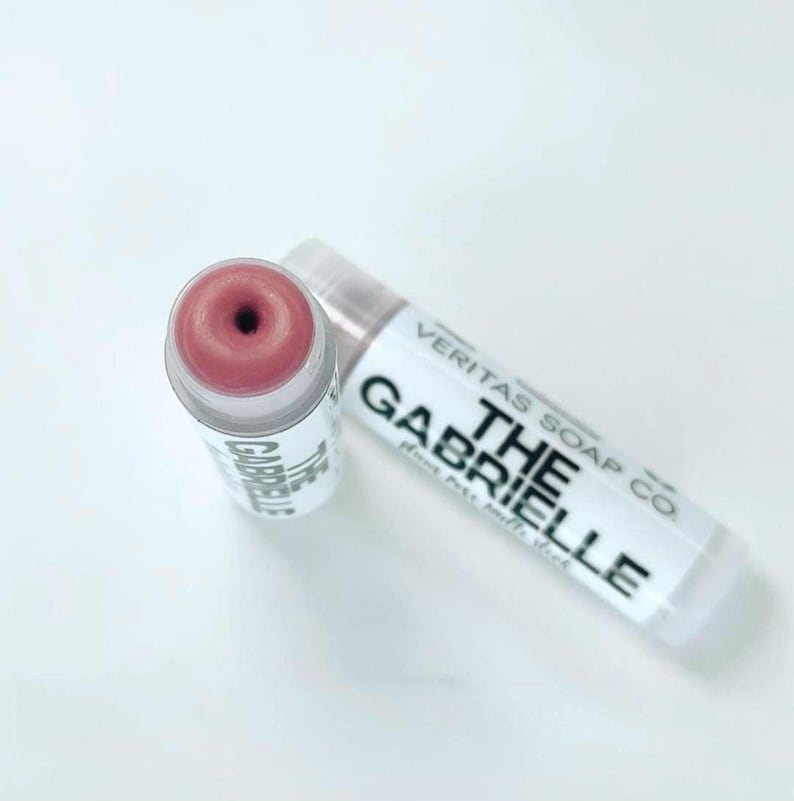 May include: Two lip balm tubes are shown on a white surface. One is open, displaying a pink balm with a dark centre. The other is closed, with the text "VERITAS SOAP CO. THE GABRIELLE" printed on the label.