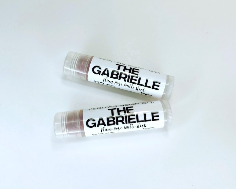 May include: Two lip balm tubes with clear caps and labels that read "THE GABRIELLE plum rose matte stick" in black text. The lip balm inside is a muted rose colour. The tubes are on a white surface.