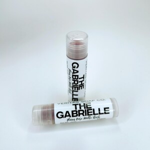 May include: Two lip balm tubes with clear caps and white labels. The labels read "THE GABRIELLE Plum Rose Multi Stick" in black text. The lip balm is a muted rose colour. The brand name "VERITAS SOAP CO." is also visible.