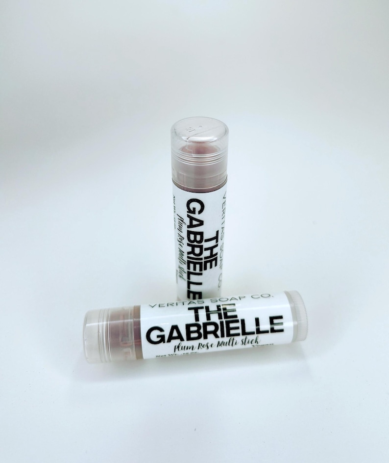 May include: Two lip balm tubes with clear caps and white labels. The labels read "THE GABRIELLE Plum Rose Multi Stick" in black text. The lip balm is a muted rose colour. The brand name "VERITAS SOAP CO." is also visible.