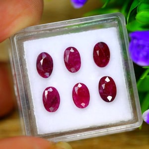 May include: Six oval-shaped ruby gemstones in a clear plastic box. The rubies are a deep red color and have a faceted cut.