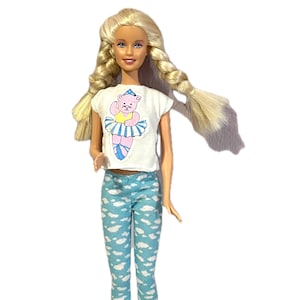 May include: A blonde doll wearing a white t-shirt with a cartoon bear in a tutu and blue leggings with a cloud pattern. The doll has long, braided hair and is standing against a white background.
