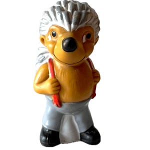 May include: A small, brown and gray plastic figurine of a hedgehog wearing a red and white backpack. The hedgehog has a white face with black eyes and a black nose.