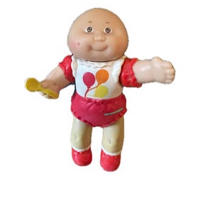 May include: A Cabbage Patch Kid doll with a bald head, wearing a red and white outfit with a bib featuring colorful balloons. The doll is holding a yellow spoon in its left hand.