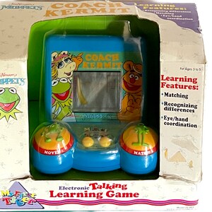 Puede incluir: Un juego de aprendizaje electrónico parlante "Coach Kermit" en caja, con personajes de los Muppets. El juego azul tiene una pantalla y dos cúpulas interactivas. La caja incluye el logotipo de los Muppets y el texto: "Electronic Talking Learning Game".