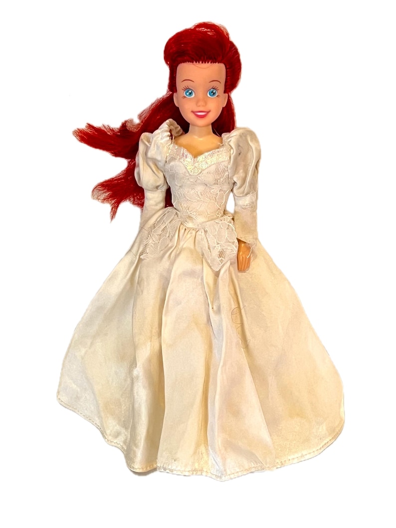 May include: A red-haired doll wearing a white satin wedding dress with lace detailing. The doll has blue eyes and a painted smile.