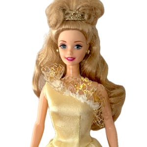 May include: A doll with long blonde hair styled in an updo with a gold headband. The doll features blue eyes, pink lips, and small gold stud earrings. It wears a sleeveless yellow satin gown with a white lace collar accented with gold sequins.
