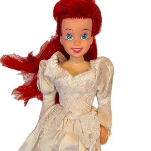 May include: A red-haired doll wearing a white lace wedding dress. The doll has blue eyes and a smiling face.