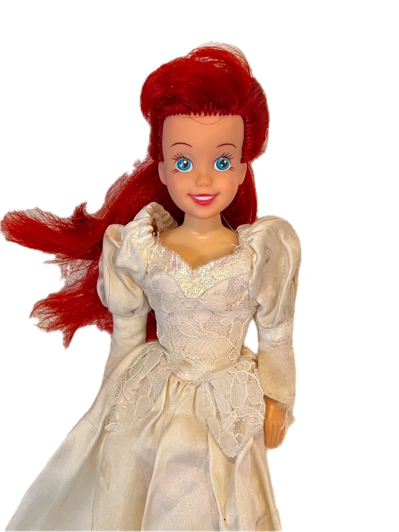 May include: A red-haired doll wearing a white lace wedding dress. The doll has blue eyes and a smiling face.