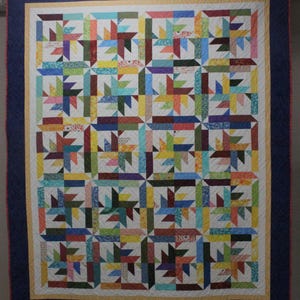 May include: A handmade quilt with a patchwork design. The quilt features a repeating pattern of colorful starbursts and geometric shapes in various colors, including red, blue, green, and yellow, set against a white background. The quilt is framed with a navy blue border.
