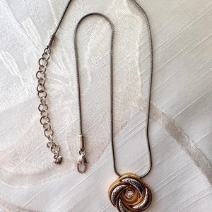 May include: A silver-toned necklace with a decorative pendant. The pendant features a gold-colored center with a clear stone and is surrounded by a swirling, patterned design. The necklace has an adjustable chain.