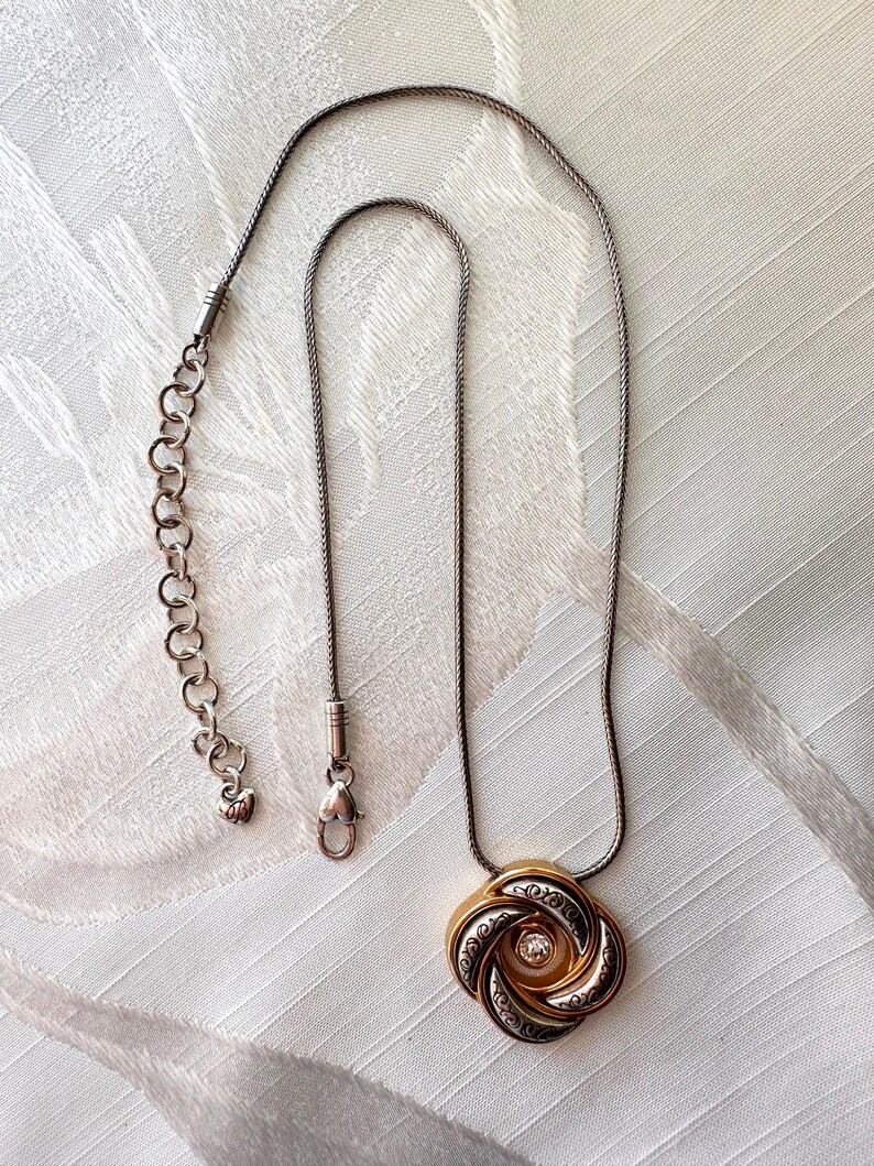 May include: A silver-toned necklace with a decorative pendant. The pendant features a gold-colored center with a clear stone and is surrounded by a swirling, patterned design. The necklace has an adjustable chain.