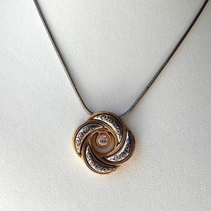 May include: A gold and silver pendant necklace with a circular design. The pendant features a gold outer layer, silver middle layer with an ornate pattern, and a small, round, pink gemstone in the center. The chain is a dark silver color.