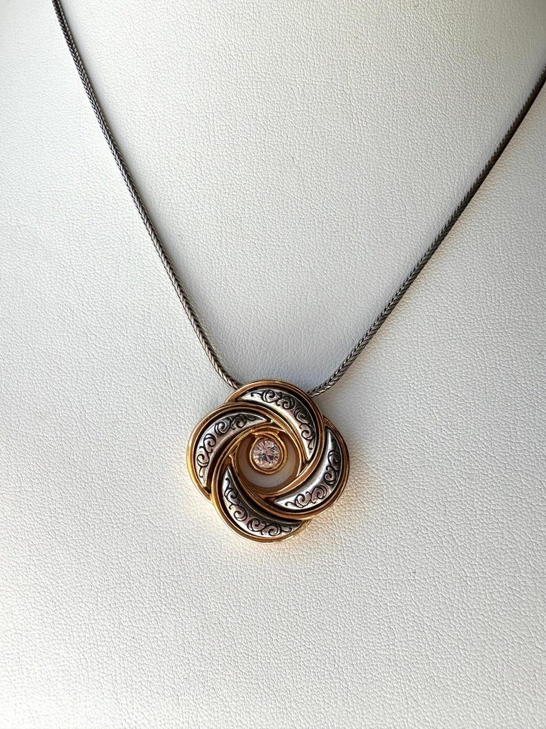 May include: A gold and silver pendant necklace with a circular design. The pendant features a gold outer layer, silver middle layer with an ornate pattern, and a small, round, pink gemstone in the center. The chain is a dark silver color.