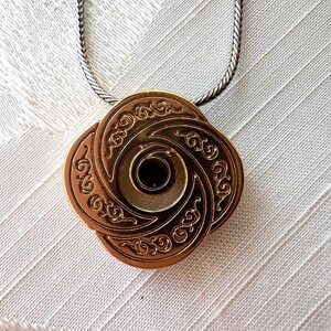 May include: A bronze-colored pendant necklace with an intricate, swirling design. The pendant is a four-petal shape with a central dark circle. The necklace chain is a silver-colored, tightly linked chain. The pendant is likely made of metal.