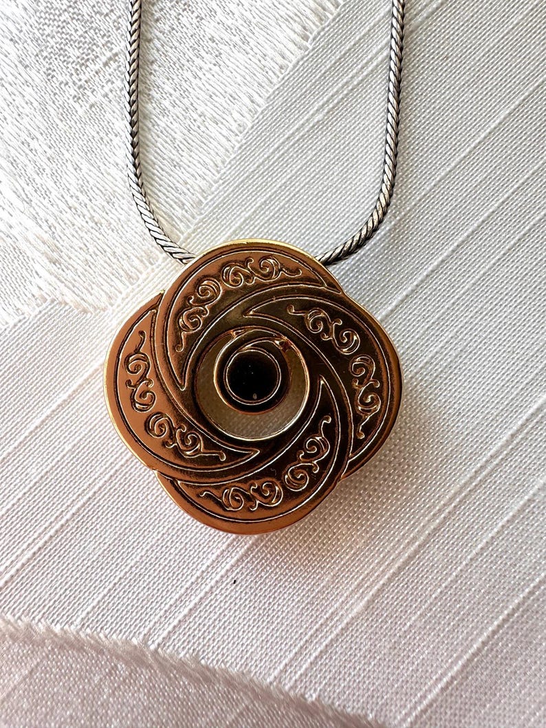 May include: A bronze-colored pendant necklace with an intricate, swirling design. The pendant is a four-petal shape with a central dark circle. The necklace chain is a silver-colored, tightly linked chain. The pendant is likely made of metal.