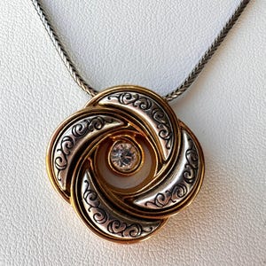 May include: A gold-toned pendant necklace with a silver-colored chain. The pendant features a swirling, four-petal design with gold and silver accents, and a clear, round gemstone in the center. The necklace is displayed on a white surface.