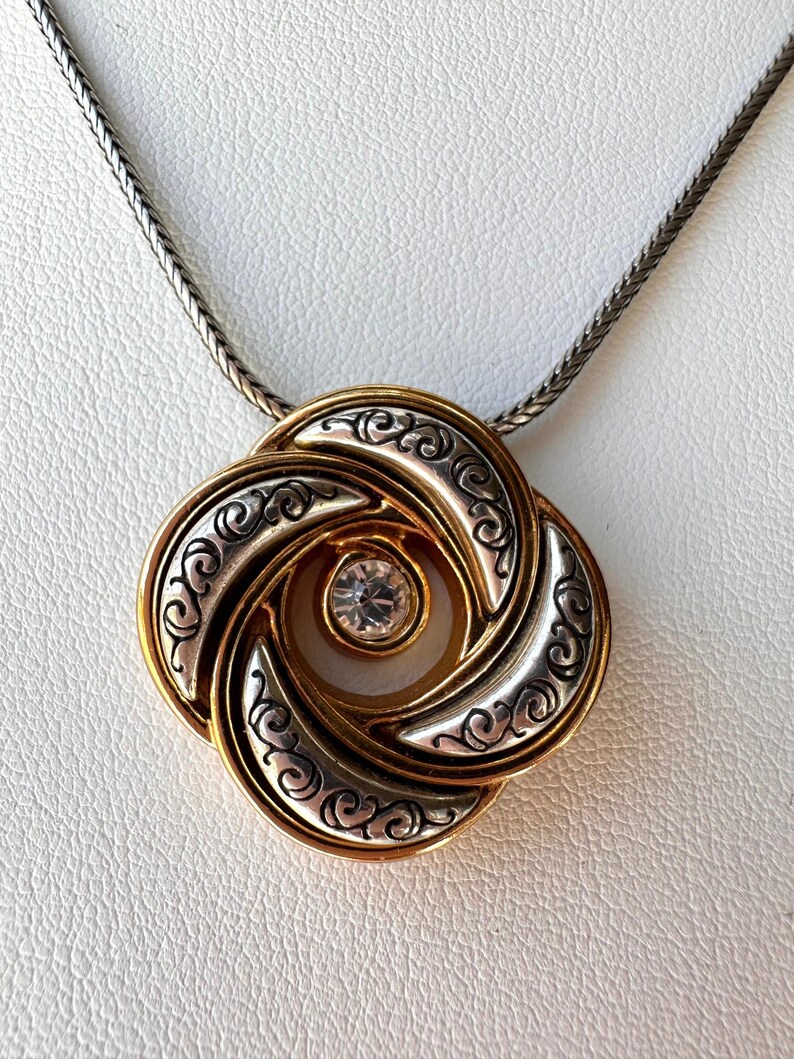May include: A gold-toned pendant necklace with a silver-colored chain. The pendant features a swirling, four-petal design with gold and silver accents, and a clear, round gemstone in the center. The necklace is displayed on a white surface.