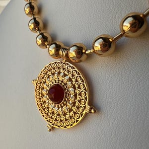 Op de afbeelding: Een goudkleurige ketting met een sierlijke hanger. De hanger heeft een centrale rode steen, omringd door kleinere heldere stenen en filigraan details. De ketting heeft een kralenketting.