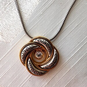 May include: A gold and silver-tone pendant necklace. The pendant features a swirling design with a central clear gemstone. The necklace has a silver-tone chain.