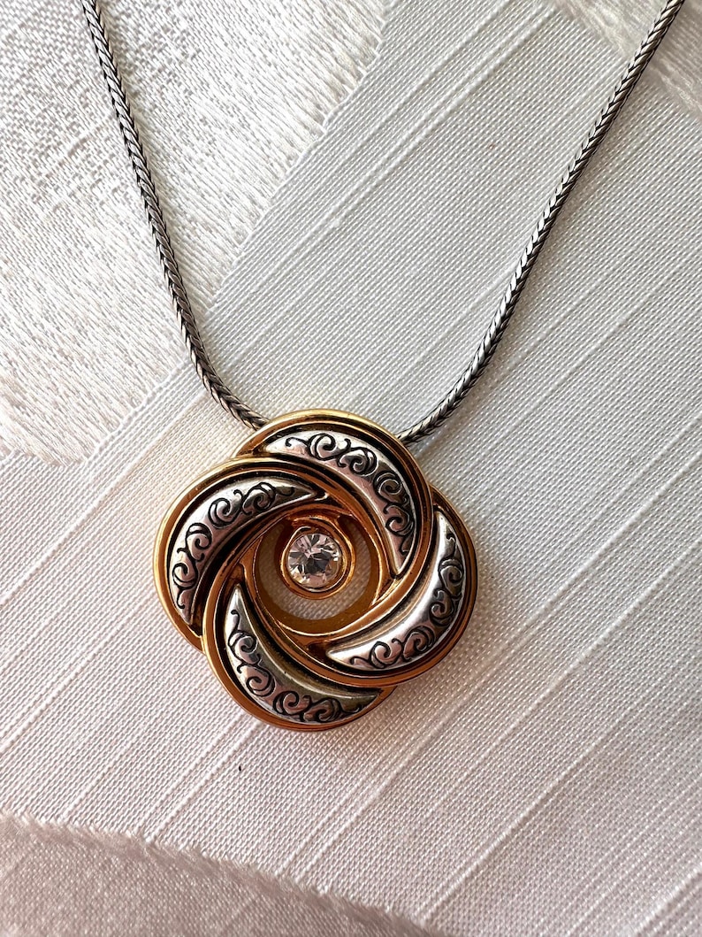 May include: A gold and silver-tone pendant necklace. The pendant features a swirling design with a central clear gemstone. The necklace has a silver-tone chain.