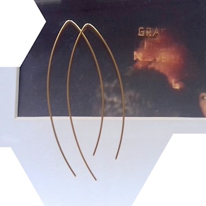 Op de afbeelding: Een paar gouden oorbellen met een eenvoudig, gebogen ontwerp. De oorbellen zijn gemaakt van dun metaal en hebben een delicate, minimalistische uitstraling.