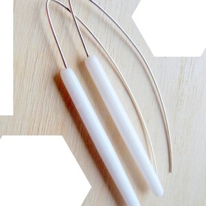 Op de afbeelding: Een paar witte, lange, cilindrische oorbellen met zilveren draadhaakjes.
