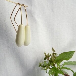 Op de afbeelding: Een paar ivoorkleurige druppelvormige oorbellen die aan goudkleurige draadhangers hangen. De oorbellen hangen tegen een witte achtergrond, met een takje groene bladeren en kleine bloemen in de rechterbenedenhoek.