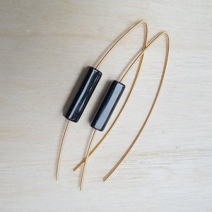 Op de afbeelding: Een paar goudkleurige draad oorbellen met een donkere, cilindrische kraal. De oorbellen hebben een eenvoudig, minimalistisch ontwerp, met de kralen gecentreerd op de draad. De oorbellen worden gepresenteerd op een lichtgekleurd houten oppervlak.