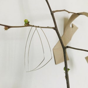 Può includere: Un paio di orecchini geometrici in argento appesi a un ramo. Gli orecchini sono fatti di filo sottile e hanno una forma triangolare.